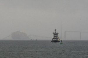A tugboat moves by a container ship as it rests against the wreckage of the Francis Scott Key Bridge